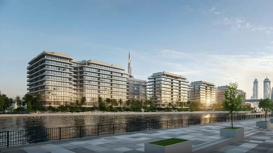 Dubai Creek Gardens seen from the opposite bank at golden hour — low-rise residential blocks along the water with a public promenade in the foreground and the Downtown Dubai skyline behind